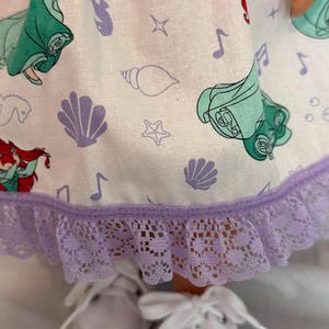 May include: A doll's dress featuring a white fabric with a repeating pattern of sea shells, musical notes, and a mermaid. The dress has a lavender lace trim and is paired with white canvas shoes.