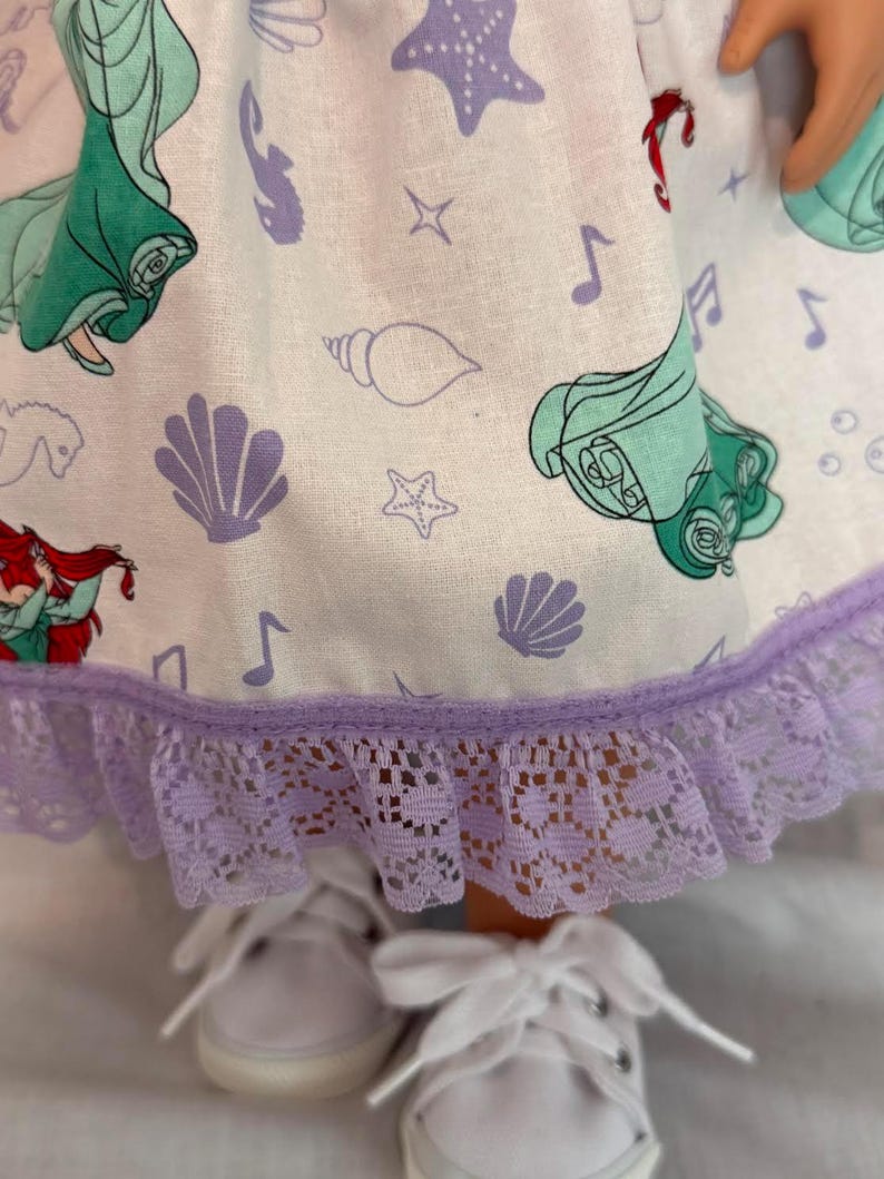 May include: A doll's dress featuring a white fabric with a repeating pattern of sea shells, musical notes, and a mermaid. The dress has a lavender lace trim and is paired with white canvas shoes.