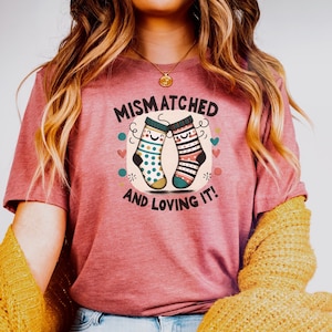 May include: A woman wearing a pink t-shirt with the text "Mismatched and Loving It!" and a pair of mismatched socks with happy faces. She is wearing blue jeans and a yellow cardigan.