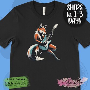 May include: A black t-shirt with a graphic of a fox playing an electric guitar. The fox is orange and white with a blue tail. The guitar is blue and white. The text "Bella+Canvas" is in green at the bottom of the shirt. The text "USA STRONG" is in white at the bottom of the shirt. The text "AIRLUME ECO-FRIENDLY" is in white at the bottom of the shirt.