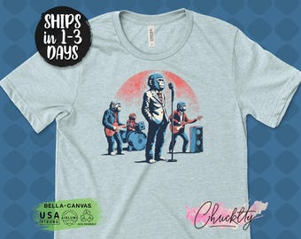 Op de afbeelding: Lichtblauw T-shirt met een band van antropomorfe apen die op het podium optreden. De illustratie toont een zanger met een microfoon, een drummer en twee gitaristen. Het ontwerp is geplaatst tegen een rode en oranje achtergrond.