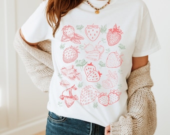 Puede incluir: Camiseta blanca con estampado de fresas rosas. Las fresas están dispuestas en un patrón en la camiseta.