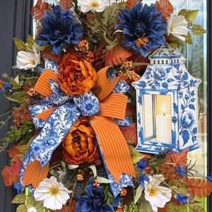 May include: A decorative wreath featuring dark blue and white flowers, orange peonies, and autumn-colored foliage. A large bow with orange stripes and blue floral patterns sits in the center, alongside a cutout of a blue and white lantern with a candle.