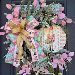 May include: A festive Christmas wreath featuring a large gold bow and a round centerpiece with a nutcracker illustration. The wreath includes pink and green accents, gold beads, and pink leaf-shaped decorations. The ribbon has a plaid pattern and nutcracker figures.