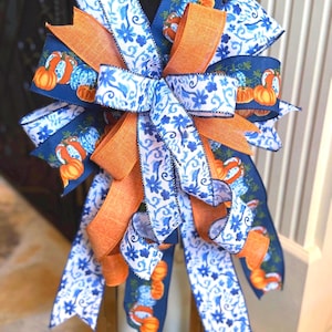 May include: A decorative bow crafted from multiple ribbons featuring a dark blue background with orange pumpkin prints, a white ribbon with blue floral patterns, and a solid orange ribbon. The bow has numerous loops and long, flowing tails.