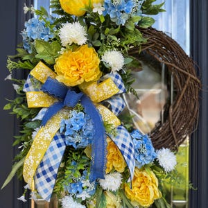 Corona de verano, hortensias azules, peonías amarillas y lazo de varias cintas.
