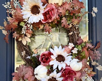 May include: A decorative wreath featuring a brown twig base adorned with white sunflowers, dark pink roses, small white pumpkins, and dried oak leaves in shades of brown and rust. The arrangement includes small white berry sprigs and green foliage.