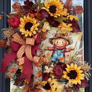 May include: A fall-themed wreath with sunflowers, pumpkins, and autumn leaves. A large bow with plaid and pumpkin-patterned ribbons is featured. A central illustration depicts a smiling scarecrow in overalls.