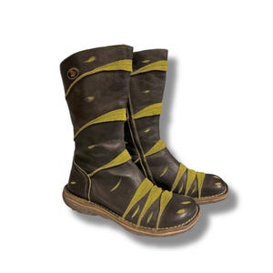 May include: A pair of dark brown leather boots with olive green accents. The boots have a mid-calf height, a side zipper, and a round toe. The olive green accents are in a zig-zag pattern across the boots. The soles are a light brown color.