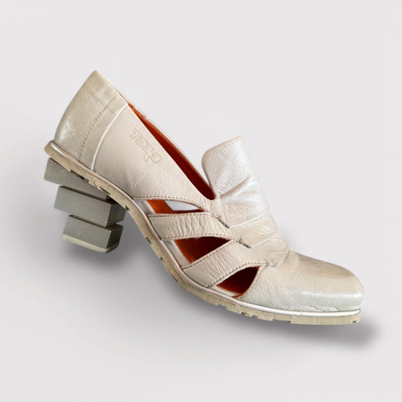 May include: White leather heeled sandals with a unique stacked heel design. The sandals have an open toe and a cut-out design on the upper.
