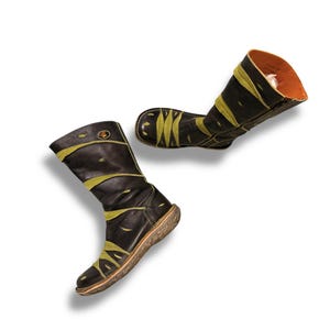 May include: A pair of black leather boots with olive green accents. The boots have a mid-calf height and a round toe. The soles are a light brown color. The interior of the boots is orange. The boots are shown against a white background.