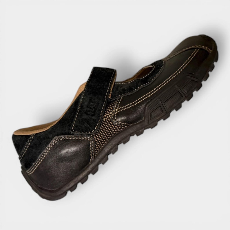 May include: Black leather shoe with a hook-and-loop strap. The shoe features a textured design on the side and a contrasting light-colored stitch. The sole is dark brown with a rugged tread. The shoe has a suede accent.