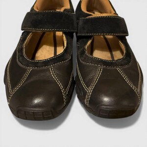 May include: Pair of black leather Mary Jane style shoes with a hook-and-loop strap closure. The shoes have a rounded toe, suede accents, and white stitching. The interior is tan, and the soles are dark brown.