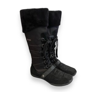 May include: Black knee-high boots with a lace-up front and a faux fur cuff. The boots have a black suede toe and heel and the brand name "Aries" is printed on the side.