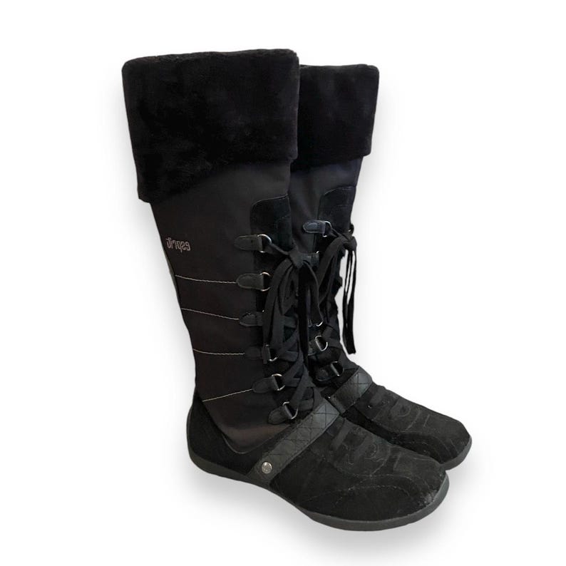 May include: Black knee-high boots with a lace-up front and a faux fur cuff. The boots have a black suede toe and heel and the brand name "Aries" is printed on the side.