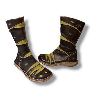 May include: Pair of black leather boots with olive green accents. The boots have a knee-high design with a rounded toe and a flat sole. Olive green straps wrap around the boots. The boots have a distressed look.