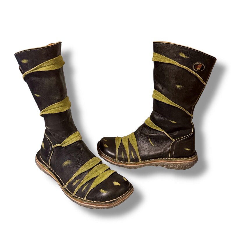 May include: Pair of black leather boots with olive green accents. The boots have a knee-high design with a rounded toe and a flat sole. Olive green straps wrap around the boots. The boots have a distressed look.