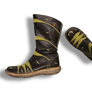 May include: A pair of dark brown leather boots with olive green accents. The boots have a mid-calf height and a textured sole. The design includes olive green leaf-like patterns across the front and sides. A small emblem is visible near the top of the boot.