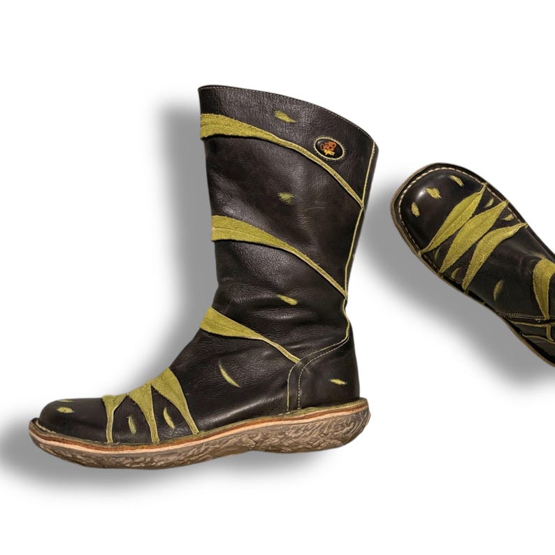 May include: A pair of dark brown leather boots with olive green accents. The boots have a mid-calf height and a textured sole. The design includes olive green leaf-like patterns across the front and sides. A small emblem is visible near the top of the boot.