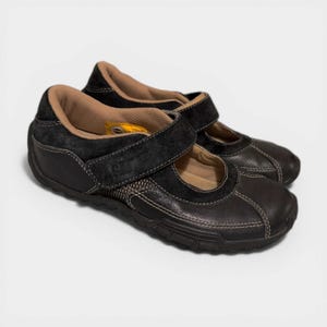 May include: A pair of black leather Mary Jane style shoes with a hook and loop closure. The shoes have a brown interior and white stitching details. The soles are black and textured.