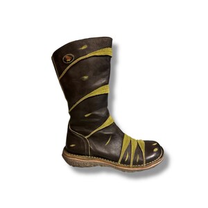 May include: A brown leather boot with olive green accents. The boot features a round toe, a mid-calf height, and a decorative design. The sole is a light brown color. A circular emblem is visible on the upper shaft.