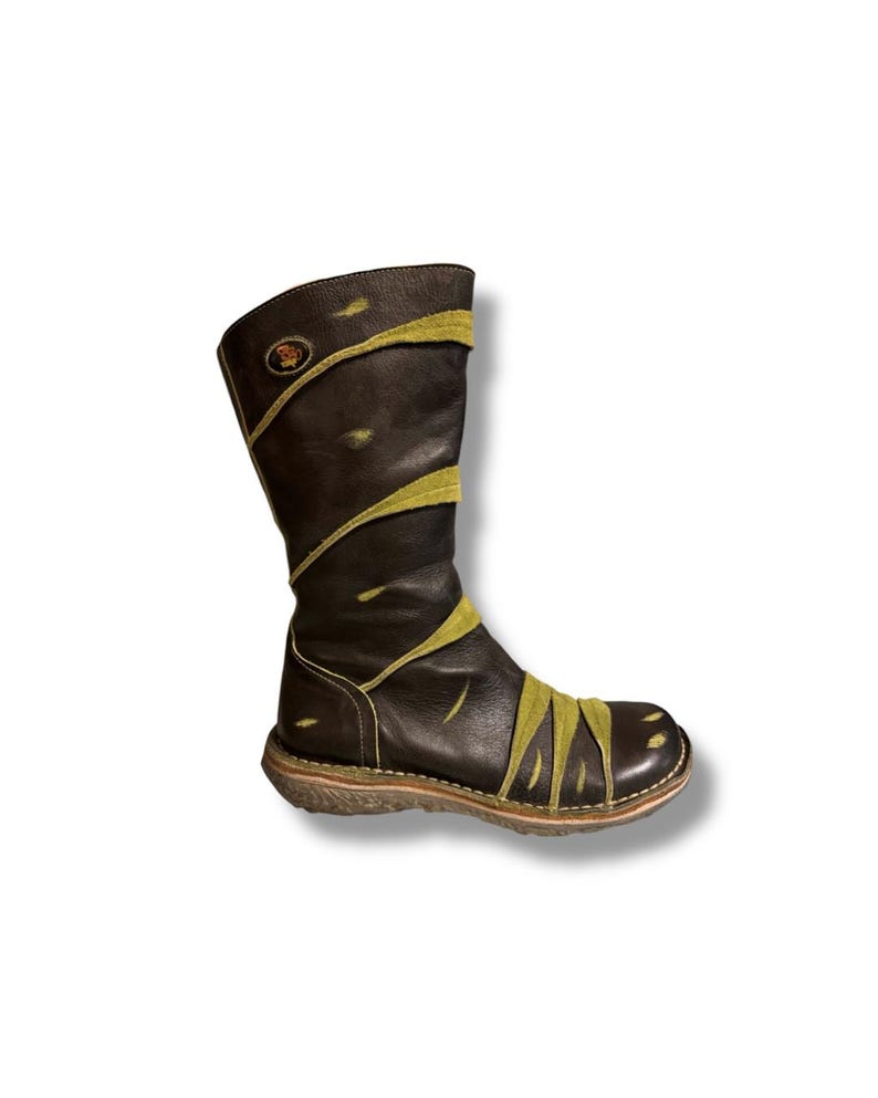 May include: A brown leather boot with olive green accents. The boot features a round toe, a mid-calf height, and a decorative design. The sole is a light brown color. A circular emblem is visible on the upper shaft.