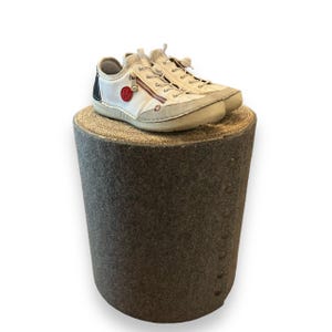 May include: A pair of white leather sneakers with a red zipper pull and a black accent on the side. The sneakers are sitting on a gray, textured cylindrical stool.