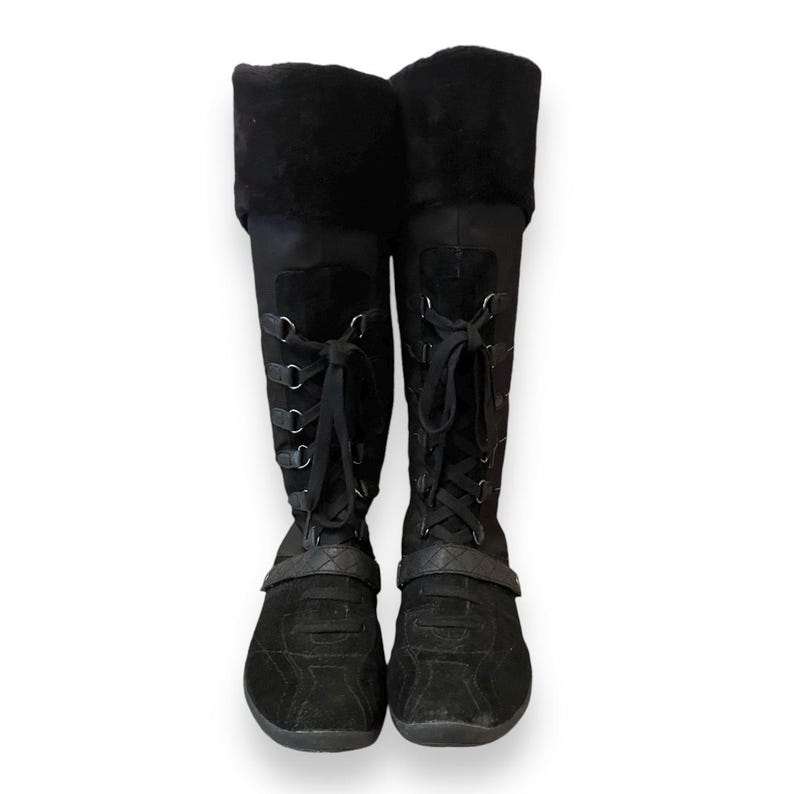 May include: Black suede knee-high boots with lace-up detailing and a black strap across the top of the foot.