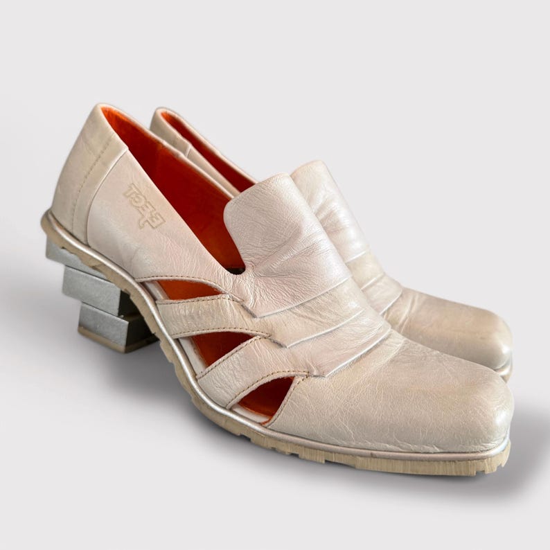 May include: White leather heeled shoes with cutouts and a silver platform. The shoes have an orange insole and the brand name "Feet" is embossed on the side.