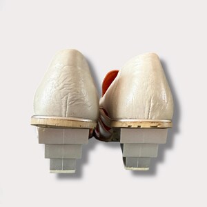 May include: A pair of white leather shoes with orange accents and stacked heels. The shoes have a pointed toe and a low heel.