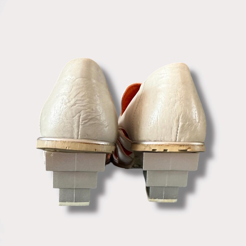 May include: A pair of white leather shoes with orange accents and stacked heels. The shoes have a pointed toe and a low heel.