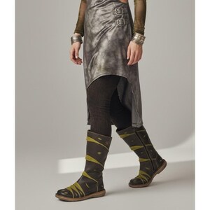 May include: A pair of dark brown leather knee-high boots with olive green accents. The boots have a round toe and a low heel. The design includes decorative stitching and straps. The boots are worn with dark leggings and a metallic-looking asymmetrical skirt.