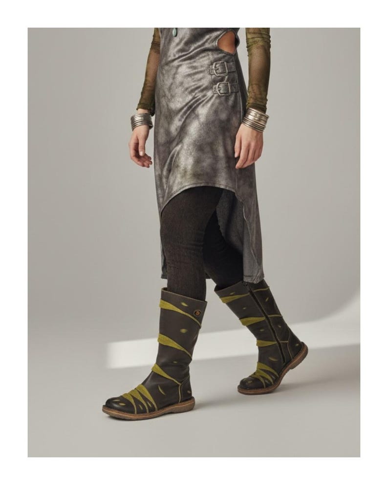 May include: A pair of dark brown leather knee-high boots with olive green accents. The boots have a round toe and a low heel. The design includes decorative stitching and straps. The boots are worn with dark leggings and a metallic-looking asymmetrical skirt.
