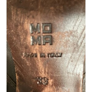 May include: Close-up of the bottom of a brown leather shoe with the text "MOMA" and "MADE IN ITALY" stamped on the sole. The size 38 is also visible.