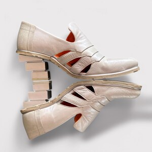 May include: A pair of white leather heeled shoes with cutouts and a stacked heel. The shoes have a pointed toe and an orange accent on the inside.