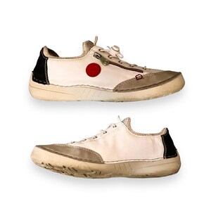 May include: Two pairs of white leather sneakers with a red circle logo and a zipper detail. The sneakers have a white rubber sole and a black trim.