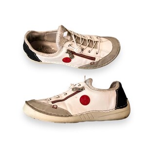 May include: White leather sneakers with red and black accents. The shoes have a zipper detail and a round red logo on the side.