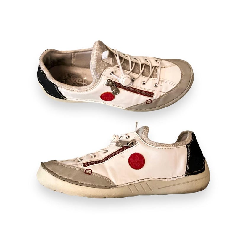May include: White leather sneakers with red and black accents. The shoes have a zipper detail and a round red logo on the side.