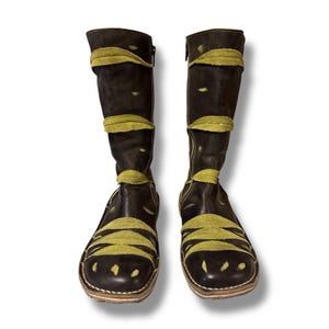 May include: A pair of knee-high black leather boots with olive green suede accents. The boots feature a zipper on the side and decorative suede bands across the shaft and toe. The soles have a stitched design.