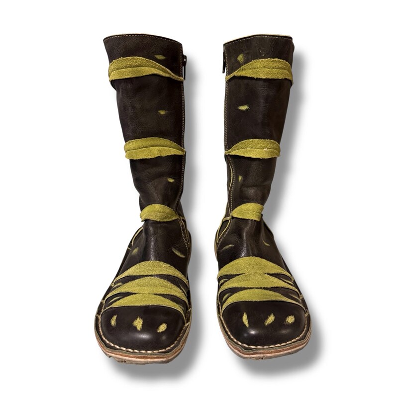 May include: A pair of knee-high black leather boots with olive green suede accents. The boots feature a zipper on the side and decorative suede bands across the shaft and toe. The soles have a stitched design.