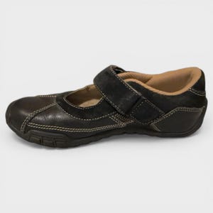 May include: Black leather Mary Jane shoe with a hook-and-loop strap. The shoe features white stitching and a tan interior. The sole is dark brown.