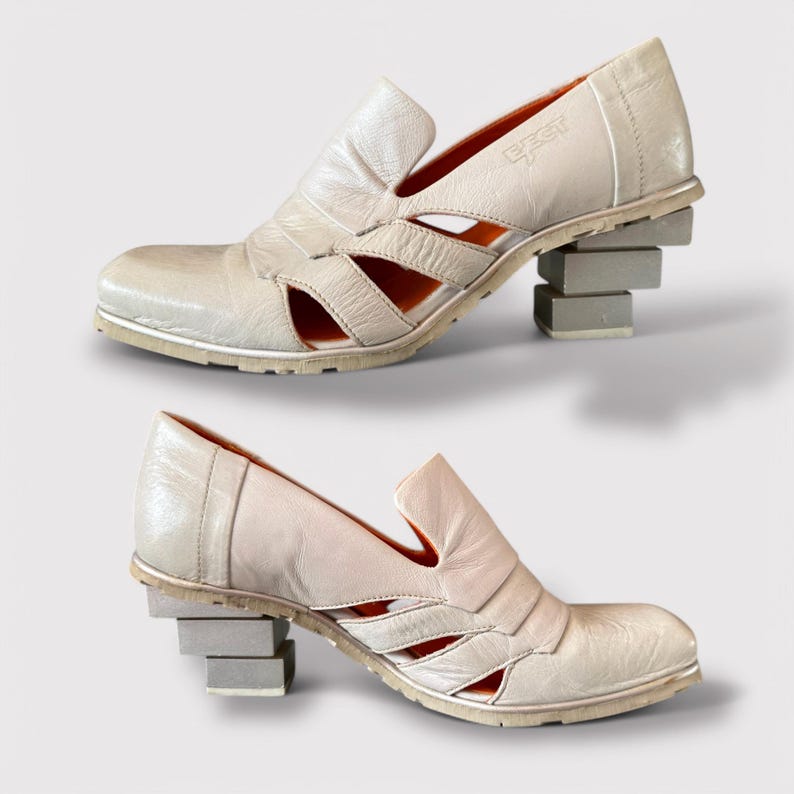 May include: A pair of white leather shoes with cutouts and a stacked block heel. The shoes have an orange lining and the brand name "gest" is visible on the top of the shoe.
