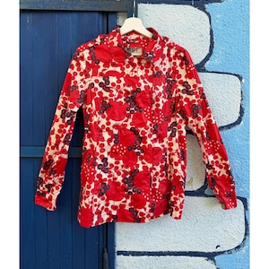 May include: A long-sleeved button-down shirt with a vibrant red and white floral pattern. The design features large red flowers and dark berries on a cream background. The shirt is hanging on a white hanger against a blue and white wall.