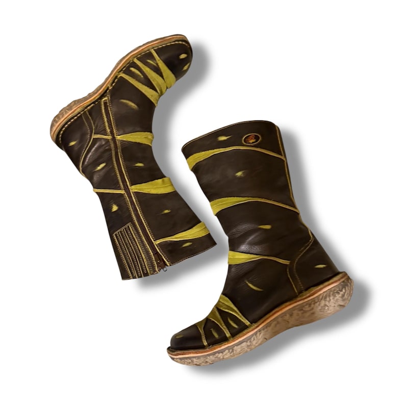 May include: A pair of brown leather boots with yellow accents. The boots have a mid-calf height, a side zipper, and a textured sole. The yellow design is abstract and covers the boots in a unique pattern.