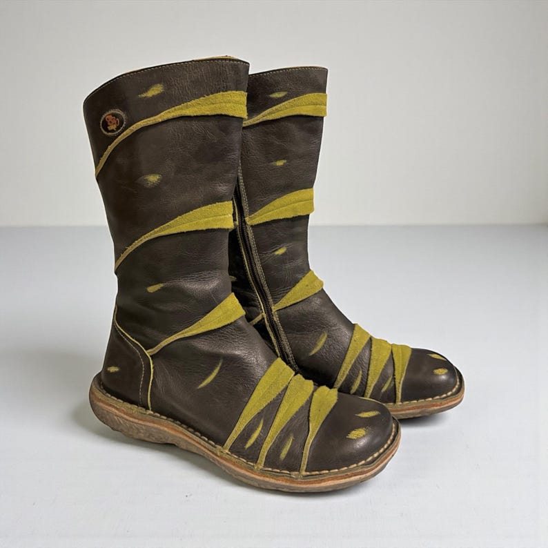 May include: A pair of brown leather boots with olive green accents. The boots have a mid-calf height, a side zipper, and a round toe. The olive green accents are in a diagonal pattern across the boots. The soles are a light brown color.