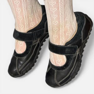 May include: Black leather Mary Jane style shoes with a velcro strap closure and white stitching. The shoes have a round toe and a thick sole. The shoes are worn with patterned cream-colored socks.