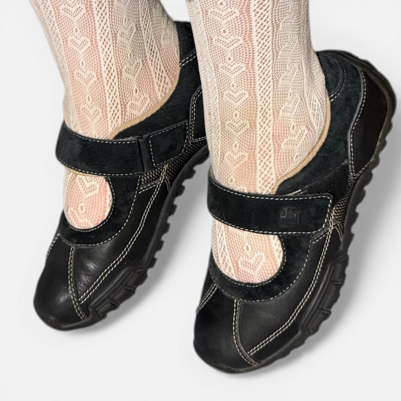 May include: Black leather Mary Jane style shoes with a velcro strap closure and white stitching. The shoes have a round toe and a thick sole. The shoes are worn with patterned cream-colored socks.