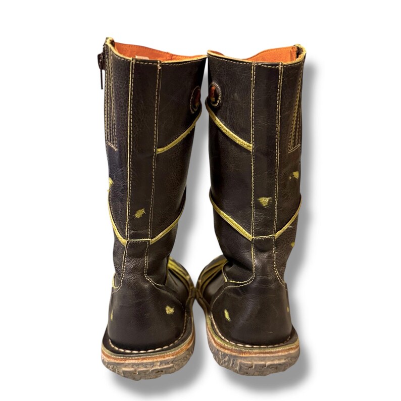 May include: A pair of brown leather boots with yellow stitching and accents. The boots have a zipper on the side and a round emblem near the top. The soles are a light brown color. The boots are knee-high.