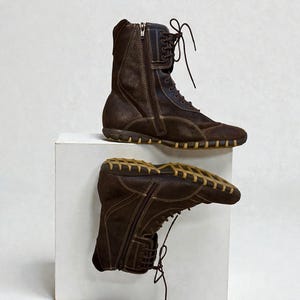 May include: A pair of brown suede boots with a side zipper and lace-up closure. The boots have a rubber sole and are displayed on a white surface. The boots appear to be new.
