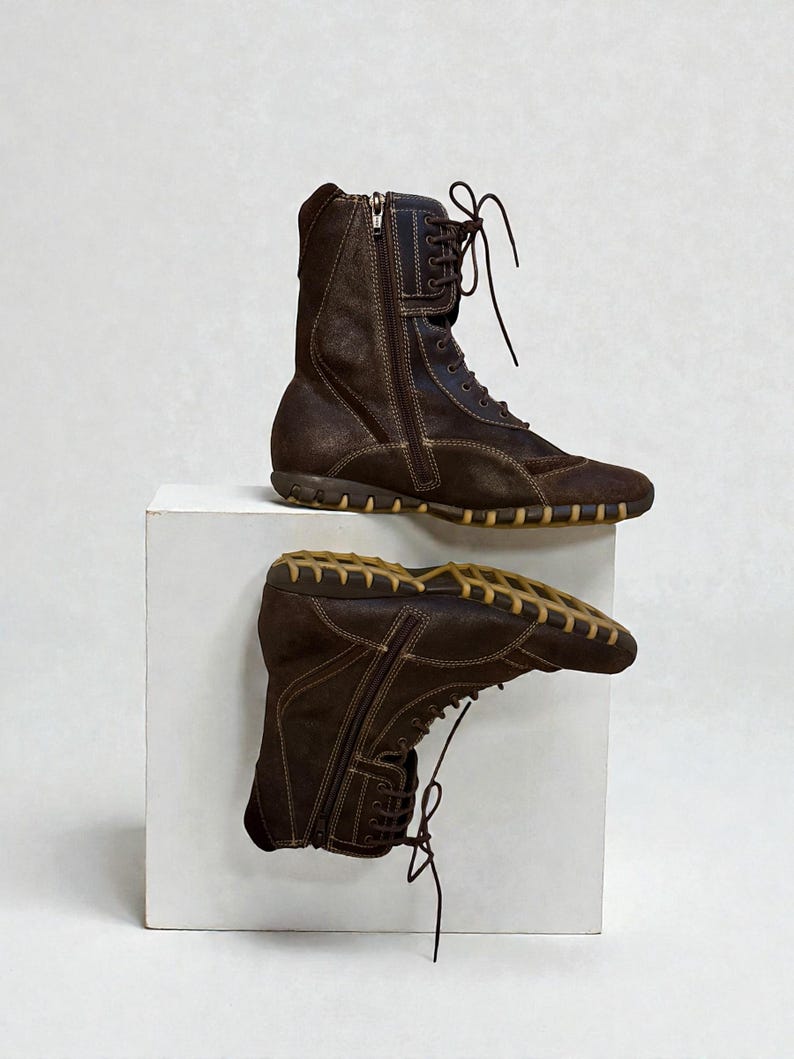 May include: A pair of brown suede boots with a side zipper and lace-up closure. The boots have a rubber sole and are displayed on a white surface. The boots appear to be new.